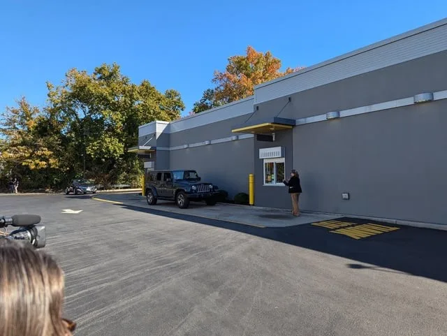 An outdoor photo of a gray commercial building with a drive-thru window. A person stands at the window, while a black Jeep…