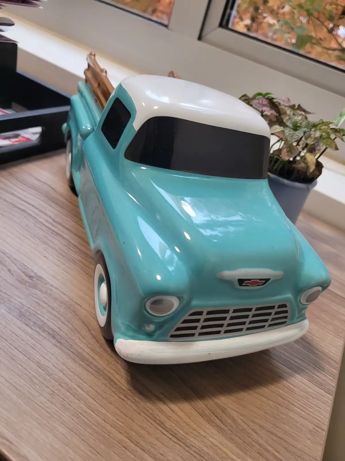 A turquoise and white ceramic toy truck with a Chevrolet emblem sits on a wooden table near a window and a potted plant. The…