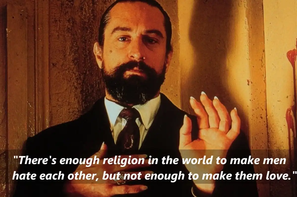 A still image of actor Robert De Niro with a beard, wearing a suit and tie, gesturing with both hands against a wooden wall…