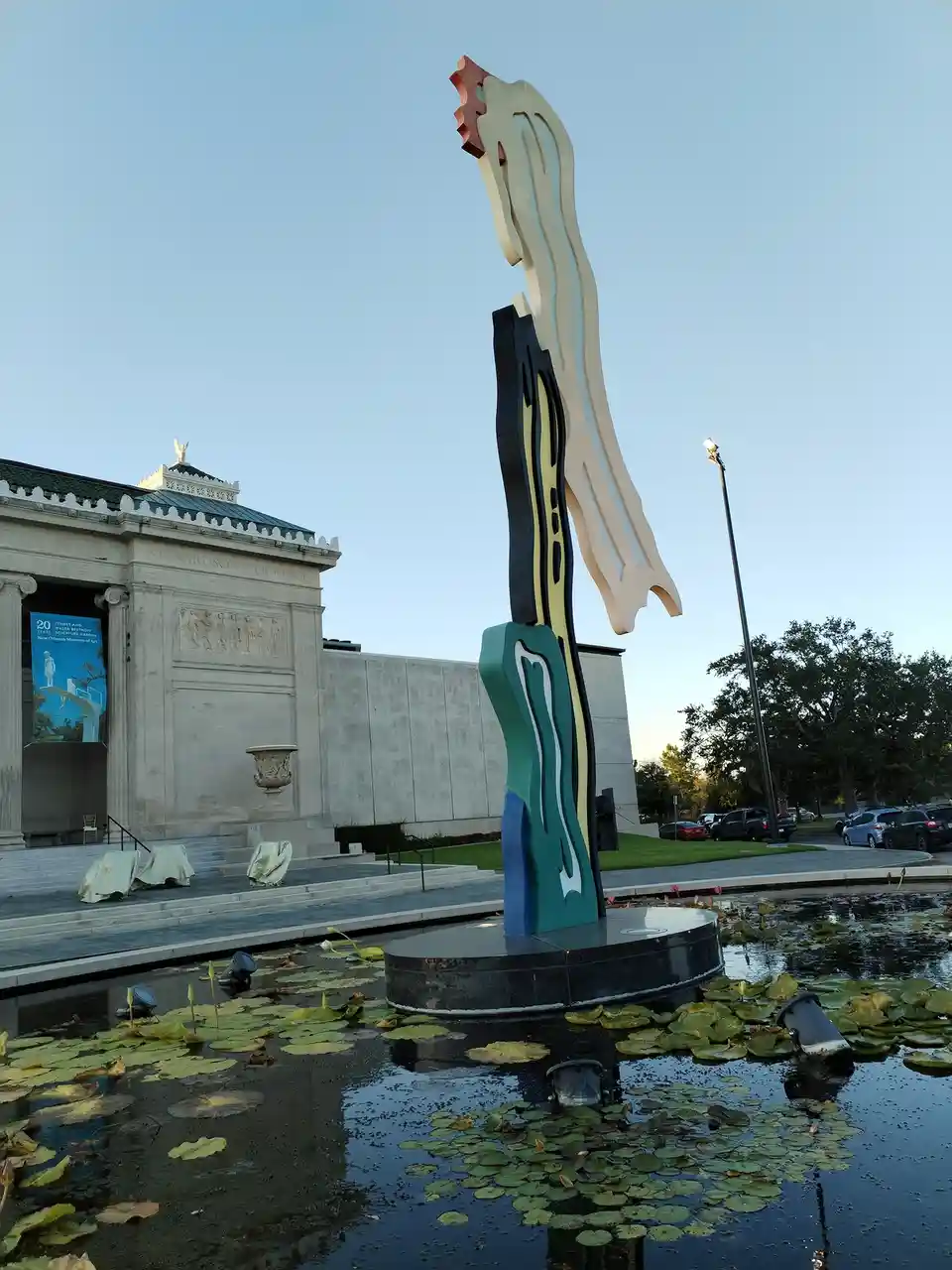 A tall, colorful abstract sculpture stands in a pond filled with lily pads, with a classical-style museum building in the…