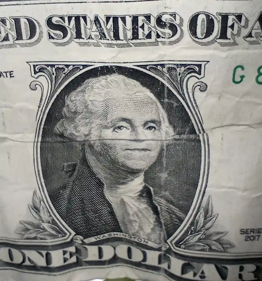 A close-up of a crumpled one-dollar bill featuring George Washington's portrait, with a horizontal crease across his face…