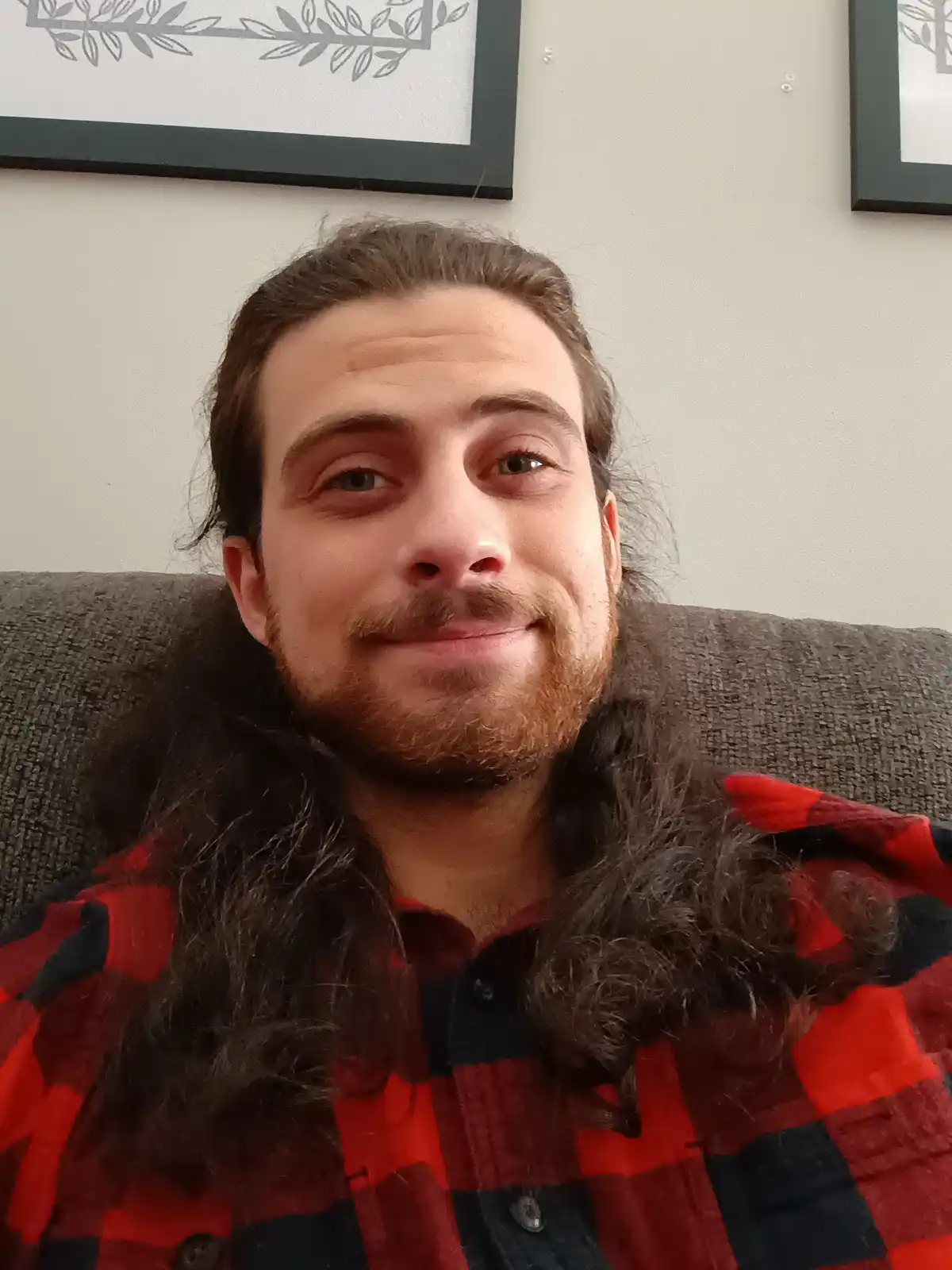 A man with long dark hair and a beard, wearing a red and black plaid shirt, sits on a gray couch and smiles slightly while…