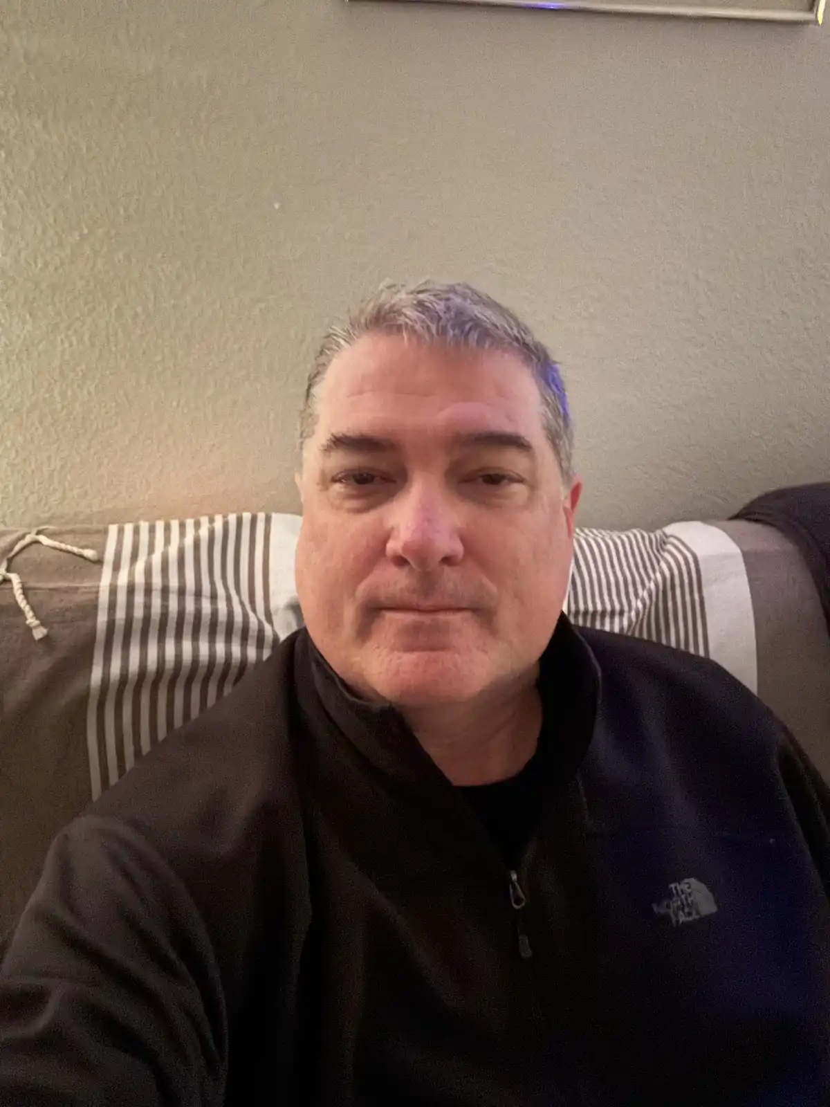 A middle-aged man with graying hair sits on a couch with a striped pillow behind him, wearing a black zip-up jacket with a…