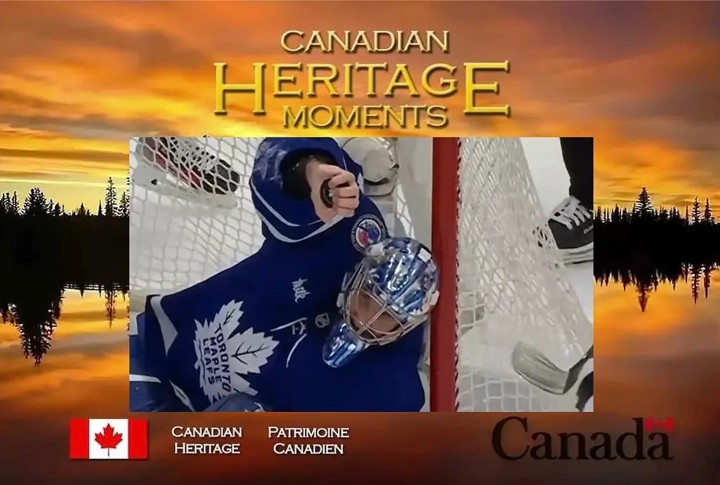 A still image of a Toronto Maple Leafs hockey goalie in a blue uniform holding a puck, framed by a sunset over water with…