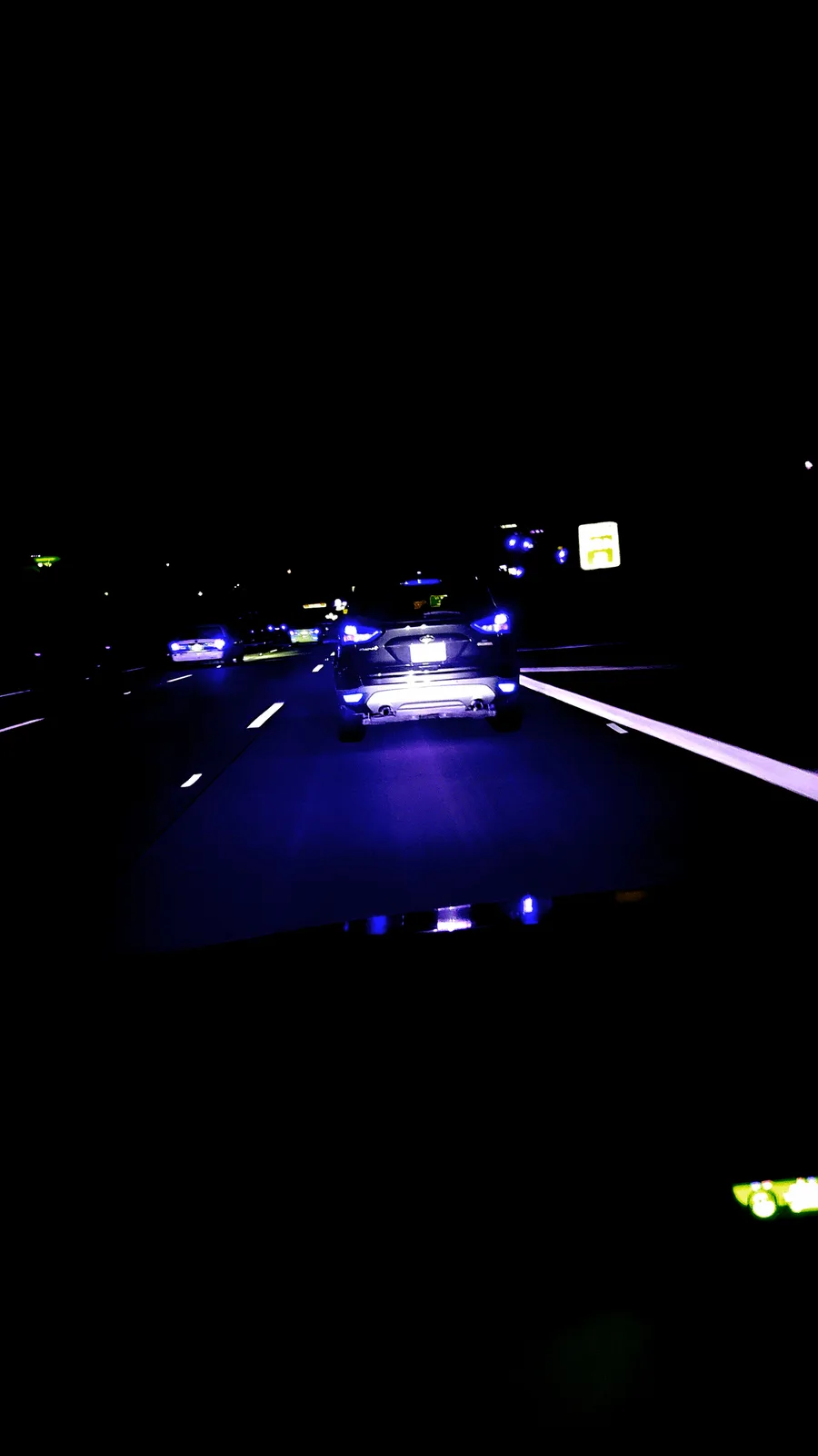 A nighttime highway photo taken from inside a car, showing the rear of a dark SUV with distinctive blue-tinted taillights…