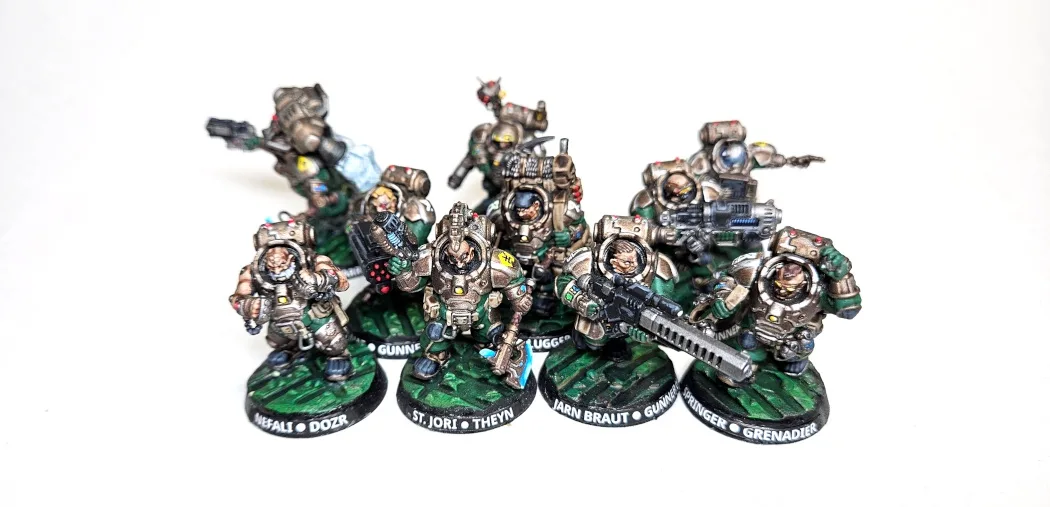 A group of eight painted, heavily armored miniature soldiers in green and grey armor, posed in combat stances on circular…