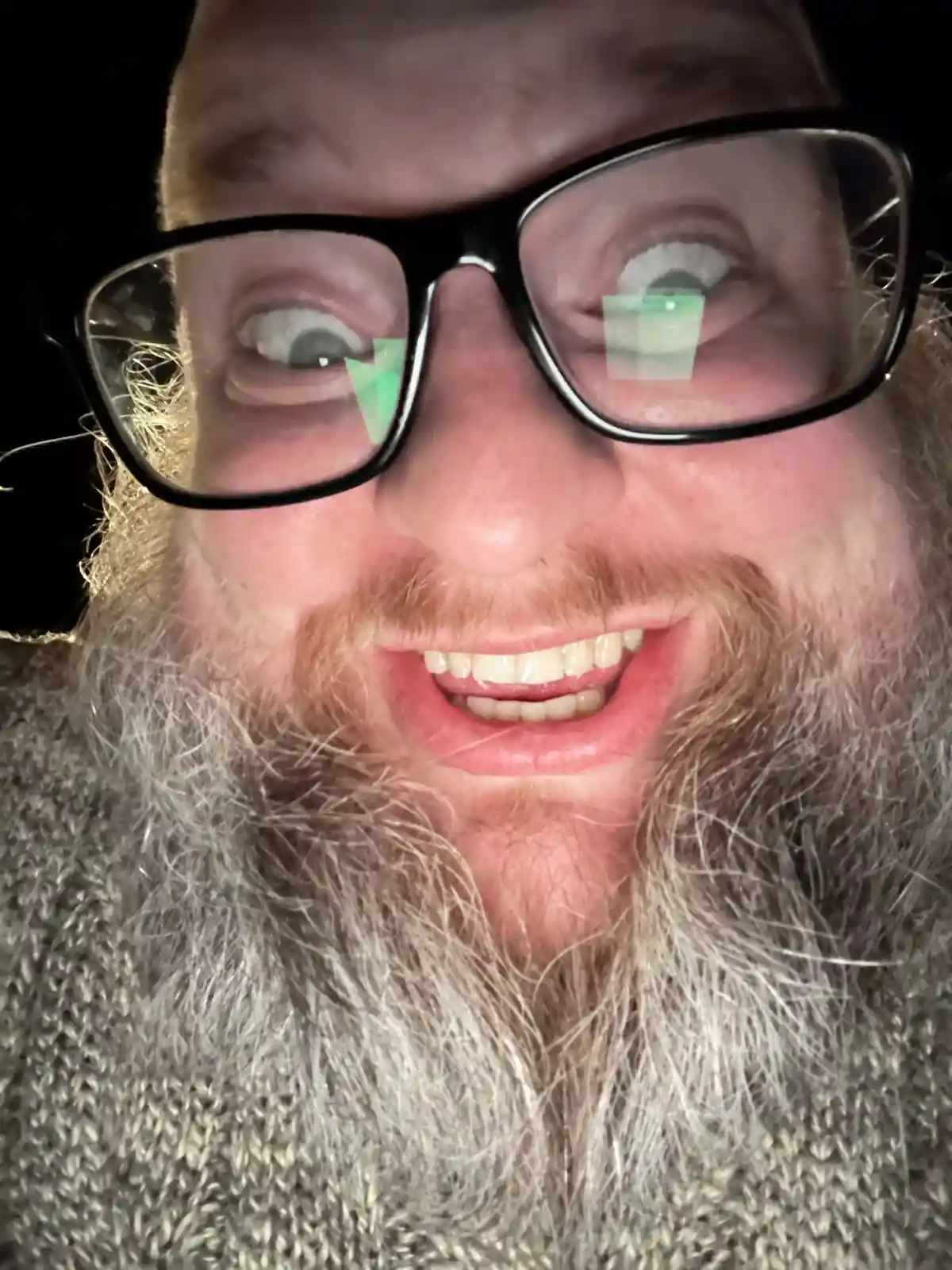 A close-up selfie of a man with a long, graying beard and black-rimmed glasses, smiling widely with his eyes wide open…