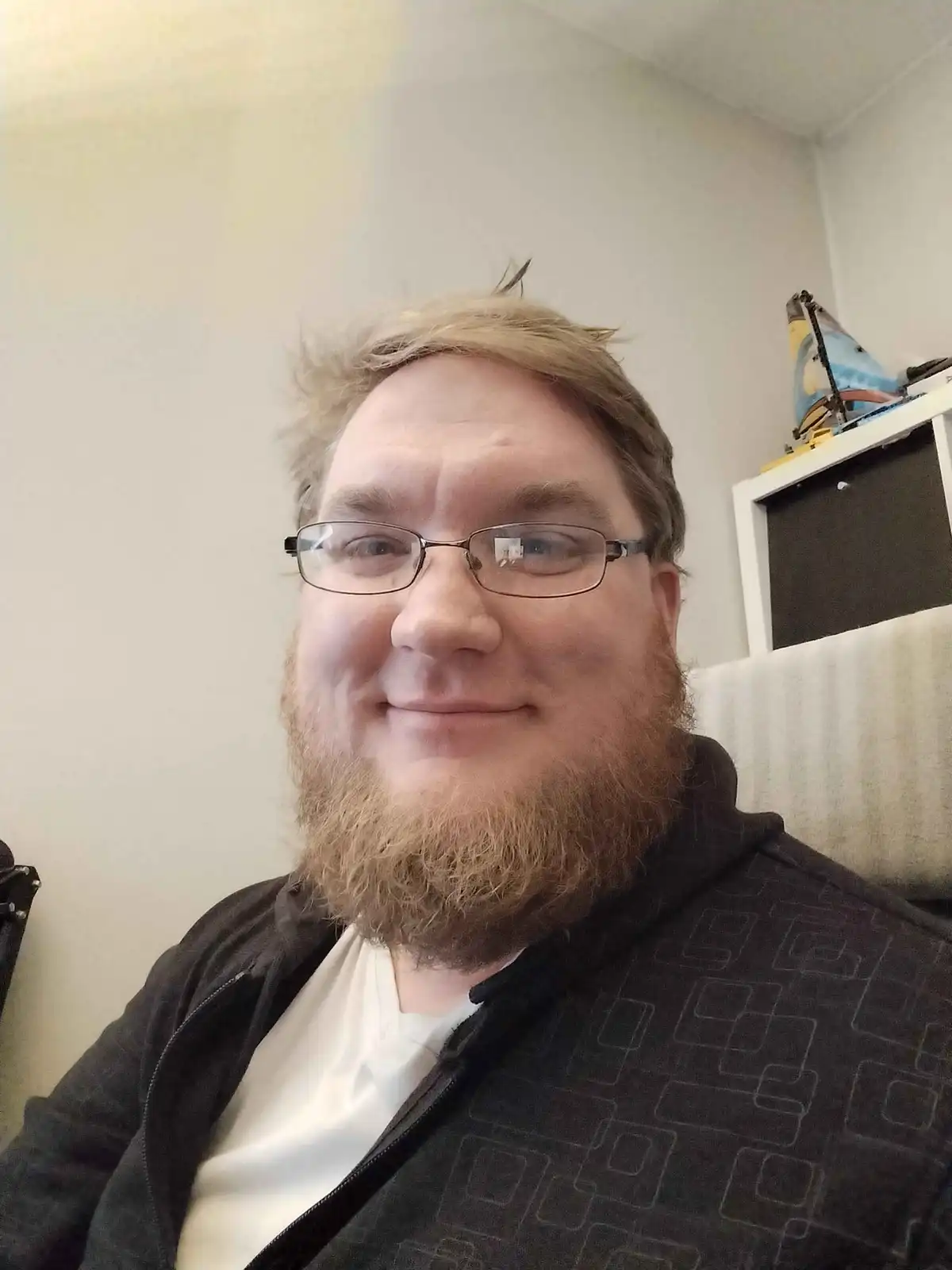 A man with glasses and a reddish-brown beard smiles at the camera while wearing a black zip-up hoodie over a white t-shirt…