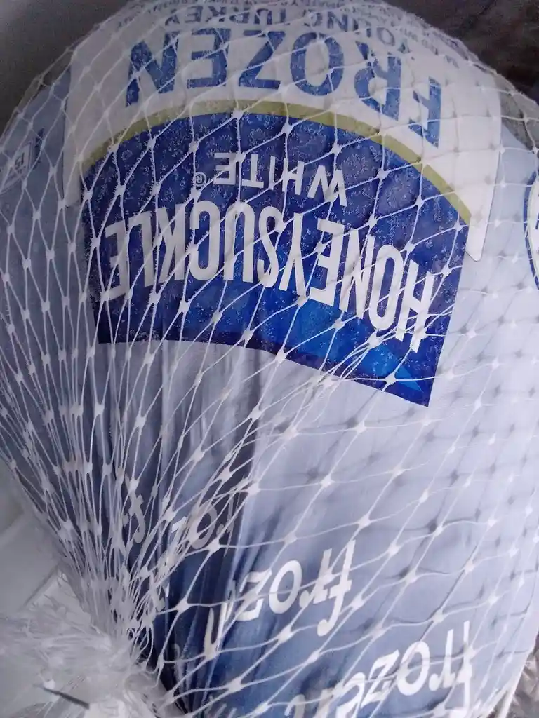 A close-up of a frozen turkey wrapped in a white plastic net, showing the label 'HONEY-SUCKLE WHITE FROZEN' partially…
