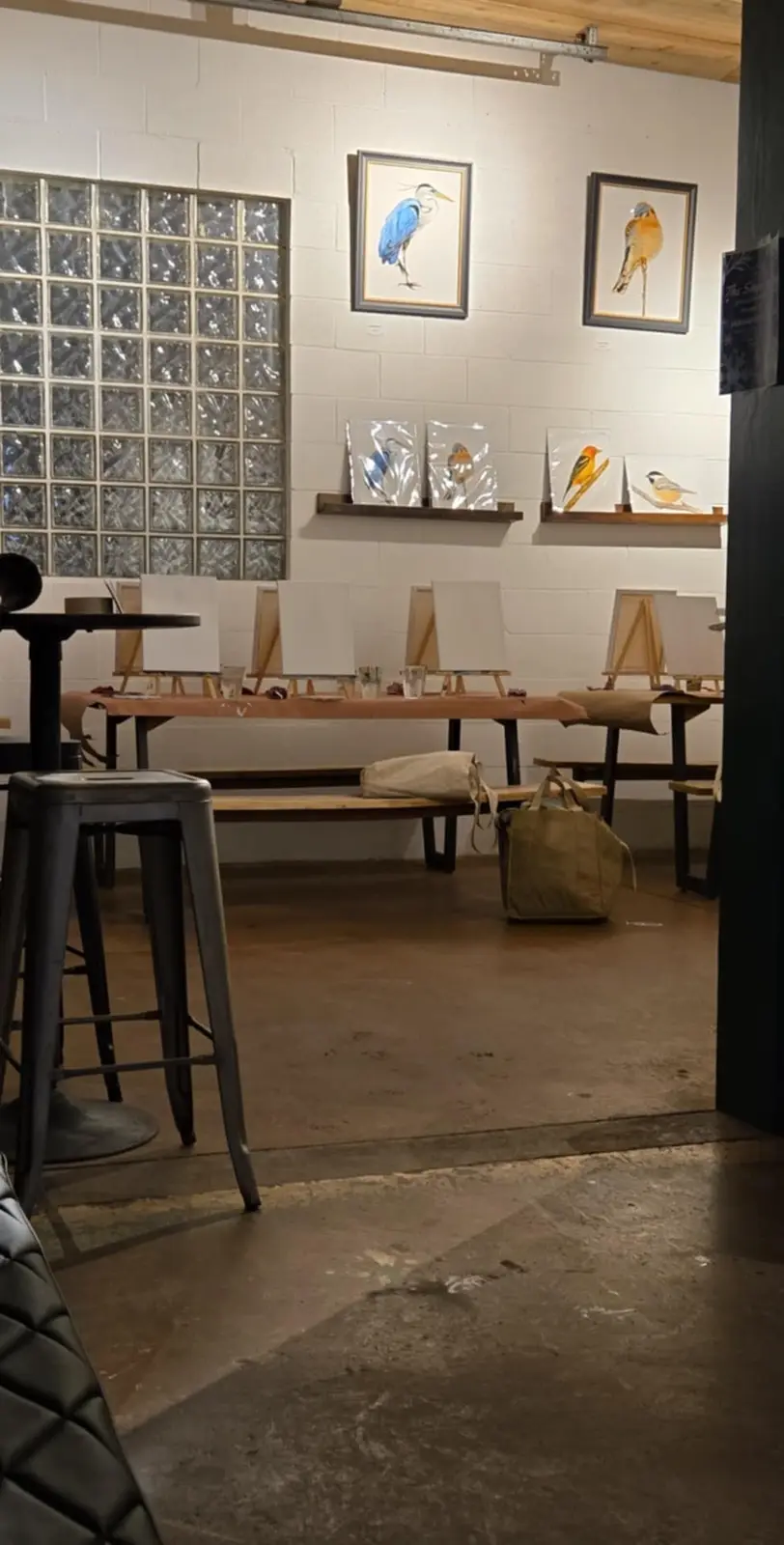 An empty art studio with blank canvases on easels, framed bird paintings on the wall, and a bag on a bench. The overlaid…