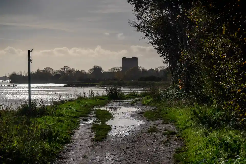 A muddy, wet path with puddles leads toward a distant castle-like structure across a body of water, framed by trees and…