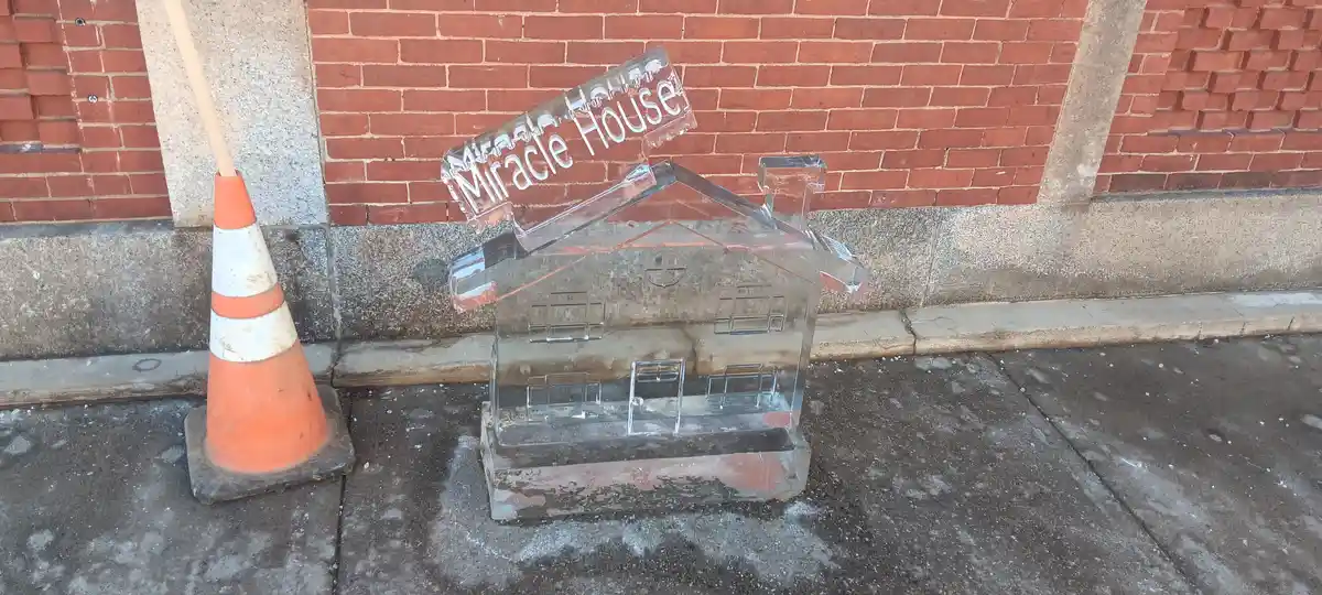 A melting ice sculpture shaped like a house with a sign reading 'Miracle House' is leaning against a red brick wall next to…