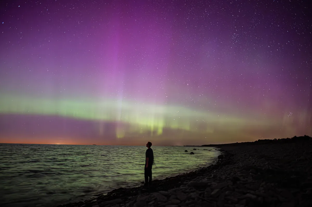 A person stands in silhouette on a rocky shoreline at night, gazing upward at a vibrant aurora borealis displaying green and…