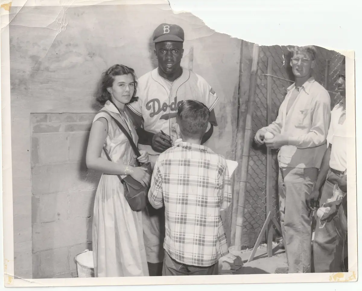 A vintage black-and-white photo shows a Black baseball player in a Dodgers uniform signing an autograph for a young boy…