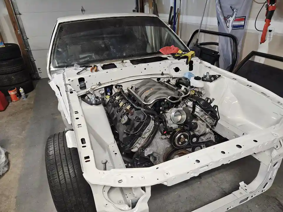 A white car's body shell sits in a garage with its hood open, revealing a fully installed engine and various tools. The…