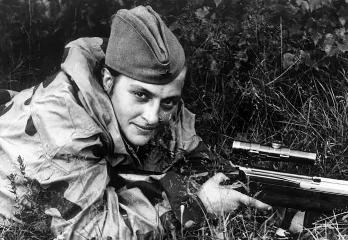 A black-and-white photo shows a man in military gear, crouched in tall grass with a rifle equipped with a scope. He wears a…