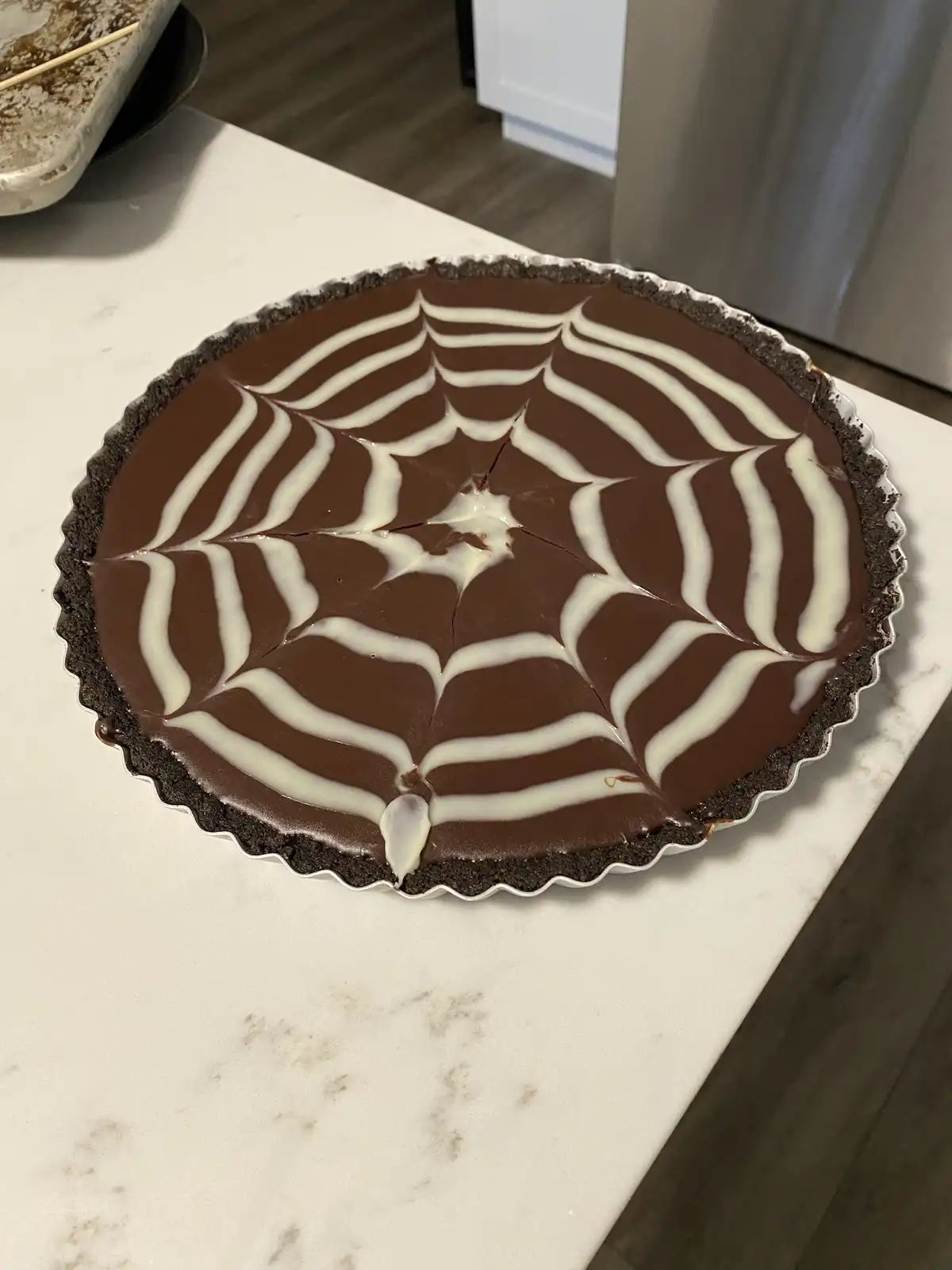 A chocolate pie with a dark crust sits on a white marble countertop, decorated with white chocolate drizzle forming a…