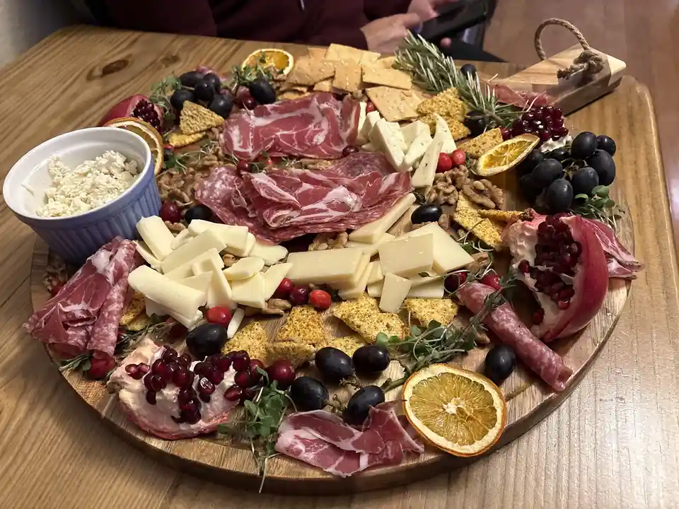 A round wooden charcuterie board is laden with an abundant spread of sliced cured meats, various cheeses, crackers, dried…