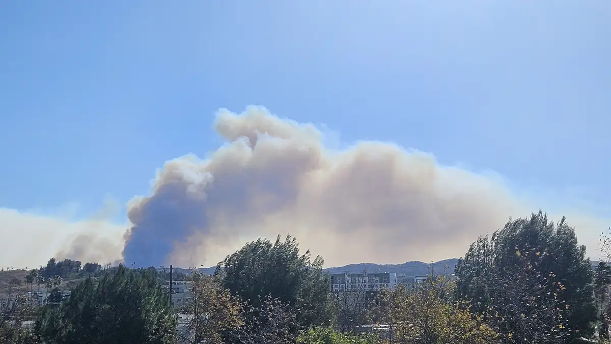 a large, billowing plume of brownish-gray smoke rises dramatically against a clear blue sky, obscuring part of distant hills…