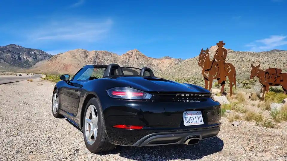 A black Porsche 718 Boxster convertible is parked on gravel beside a desert highway, with a rusty metal sculpture of a…