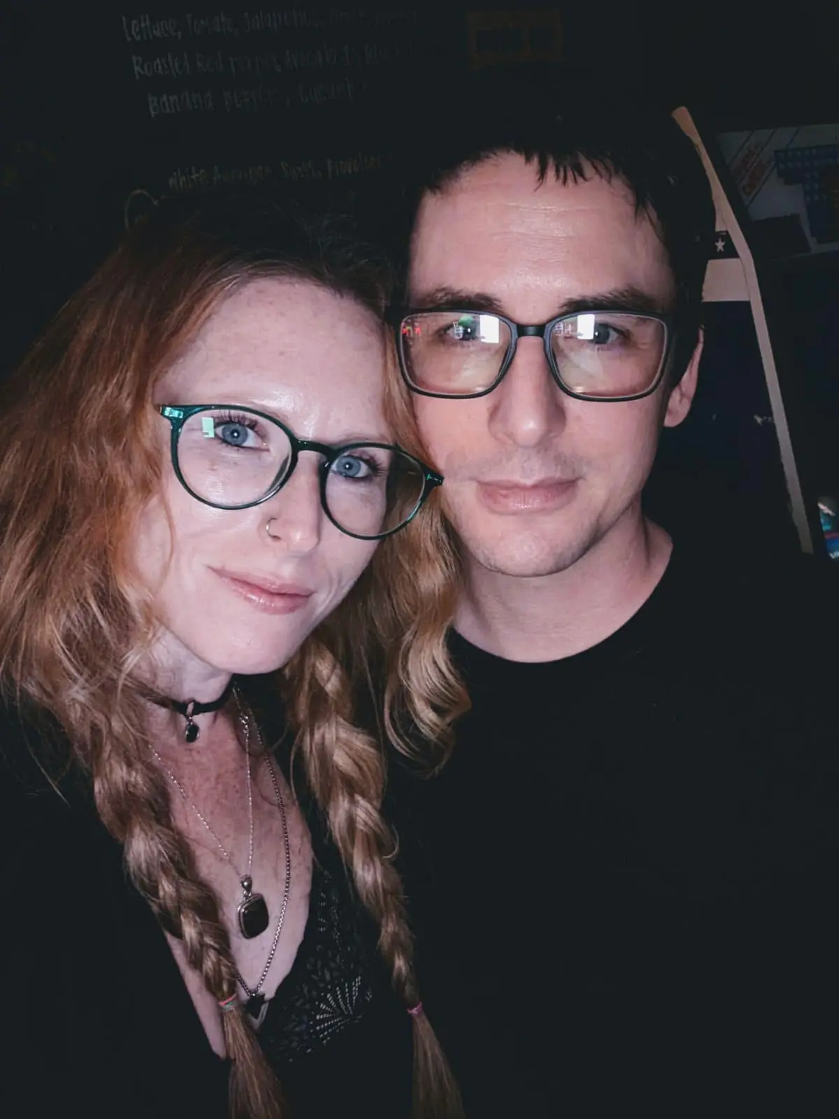 A selfie of a man and a woman with glasses, both looking at the camera. The woman has red hair in braids and a nose ring…