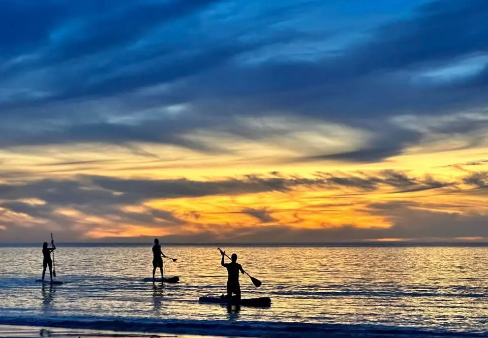 Three silhouetted people stand on paddleboards in calm water during a vibrant sunset with orange and blue clouds. The person…