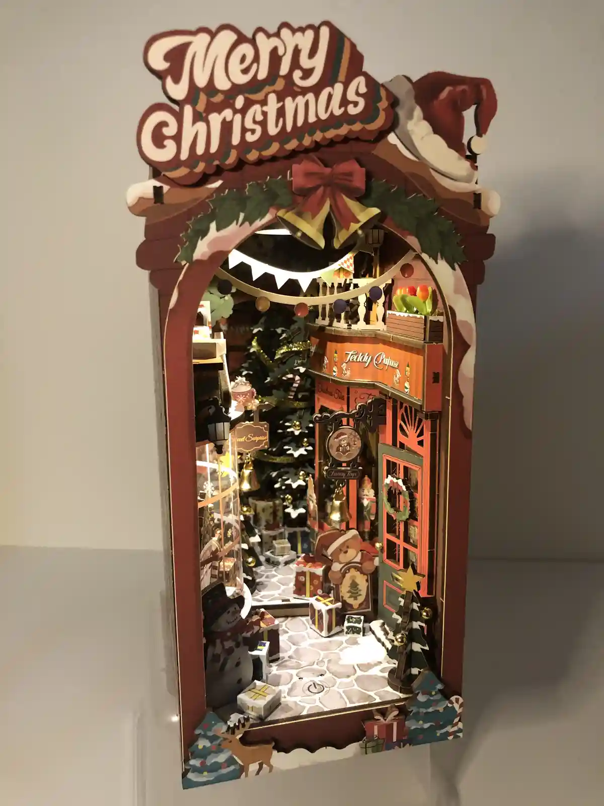 A detailed, illuminated Christmas-themed diorama with a red archway, a 'Merry Christmas' sign, and a Santa hat on top…