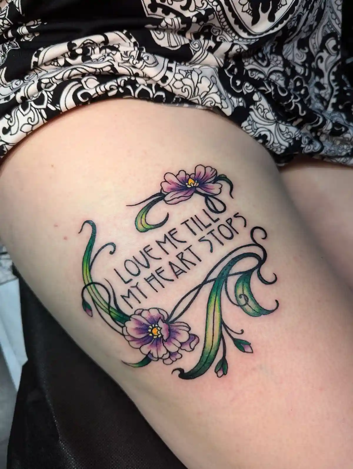 A close-up photo shows a colorful tattoo on a person's thigh, featuring two purple flowers with green stems and leaves…