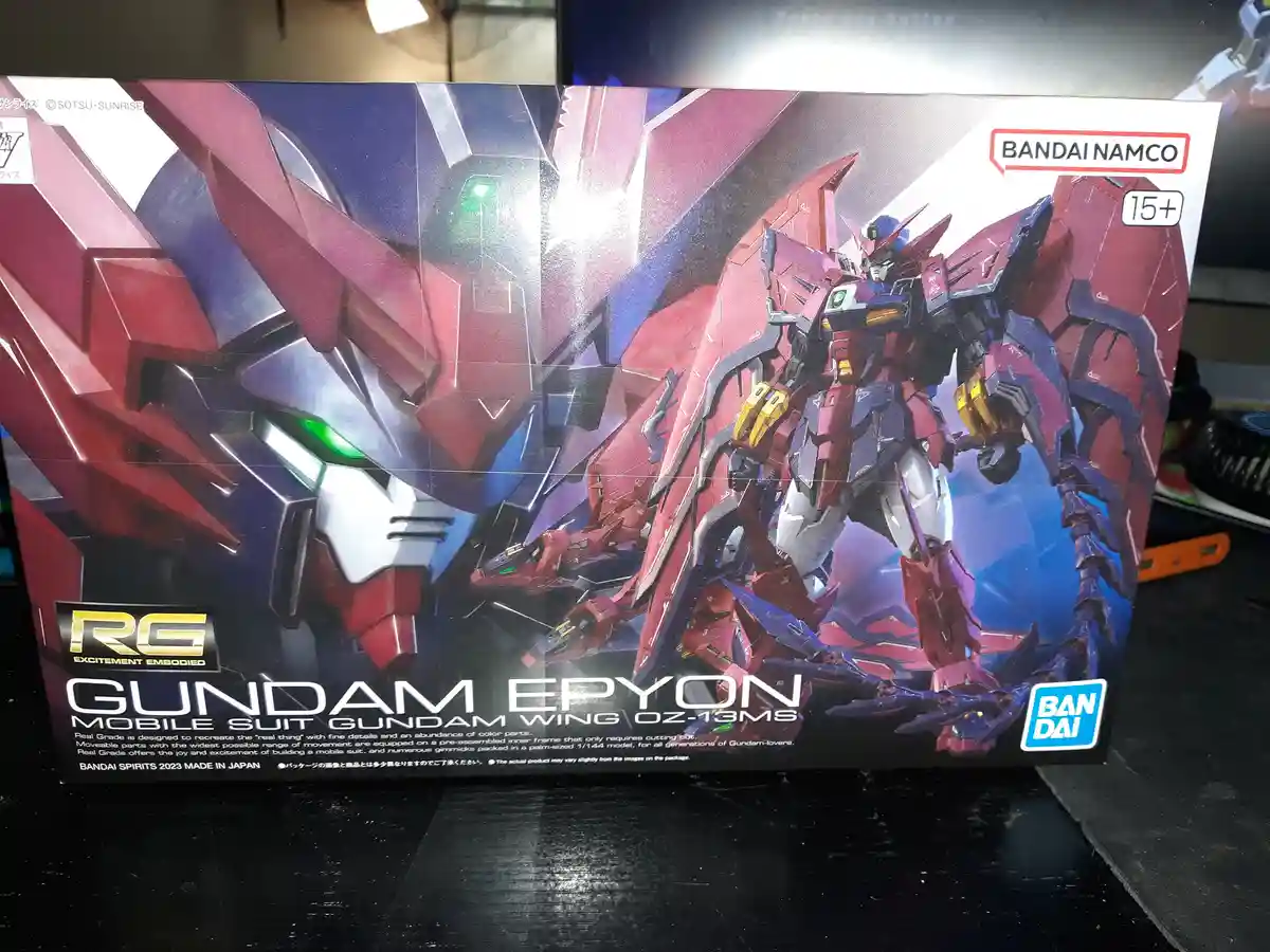 A photo of a Bandai Real Grade Gundam Epyon model kit box, featuring a red and blue mobile suit with green eyes, set against…