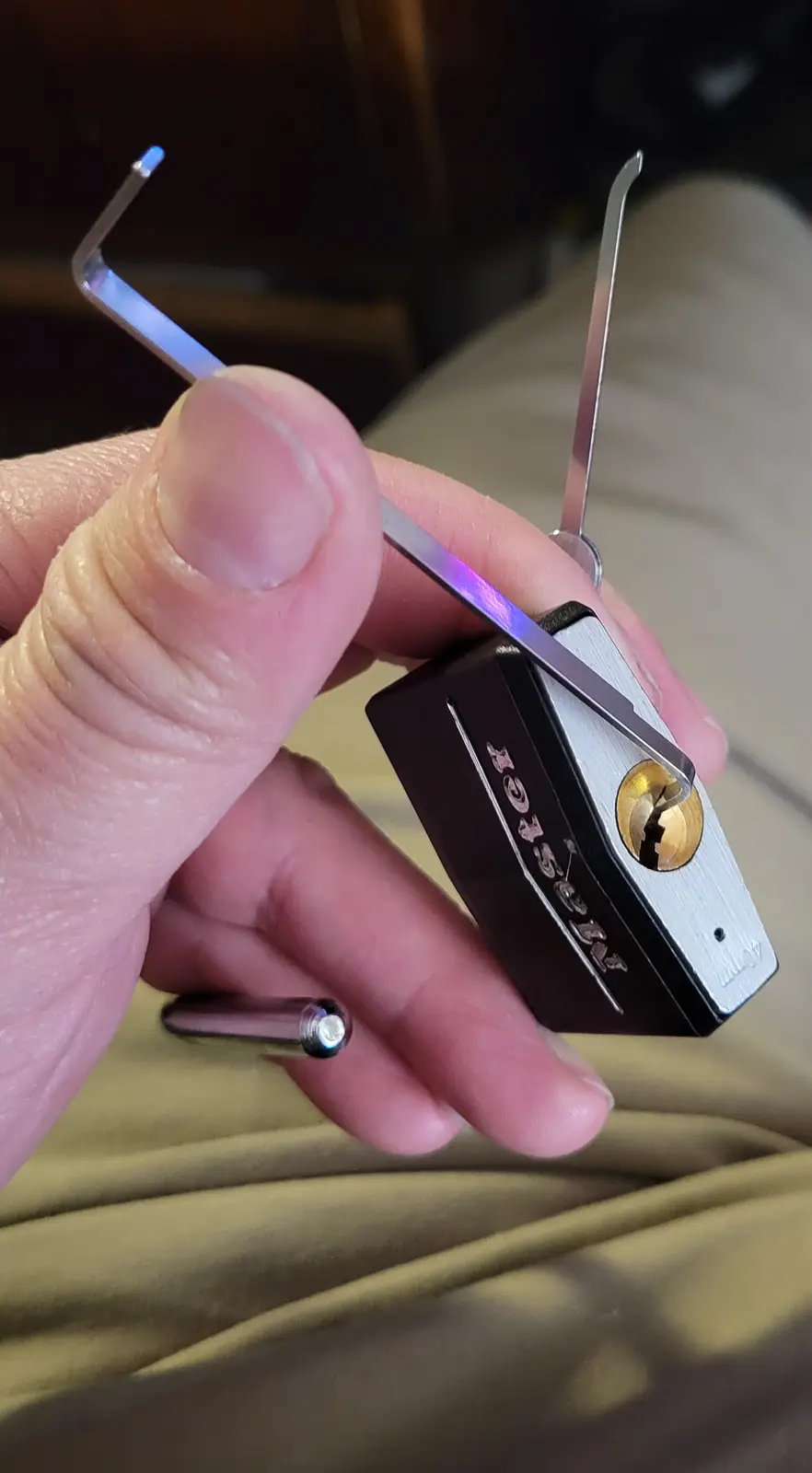 A hand holds a black and silver padlock with a broken keyhole, using a lock-picking tool inserted into the mechanism. The…