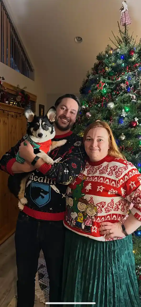 A man and woman stand in front of a decorated Christmas tree, both wearing festive, overly patterned sweaters. The man holds…