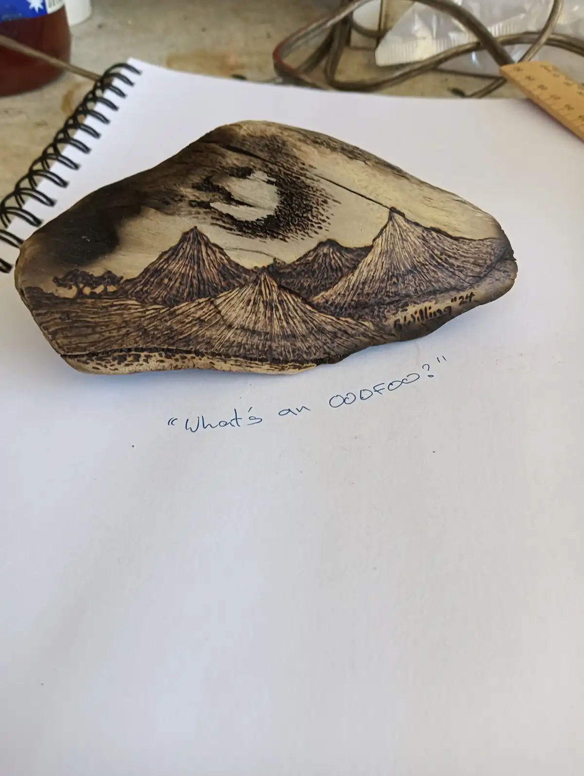 A piece of wood with a detailed pyrography landscape of mountains and sky rests on a white surface, with handwritten text…