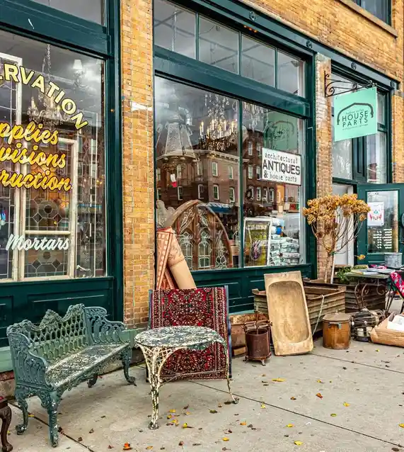 A storefront with large windows displays vintage architectural antiques and house parts, including ornate metal furniture…
