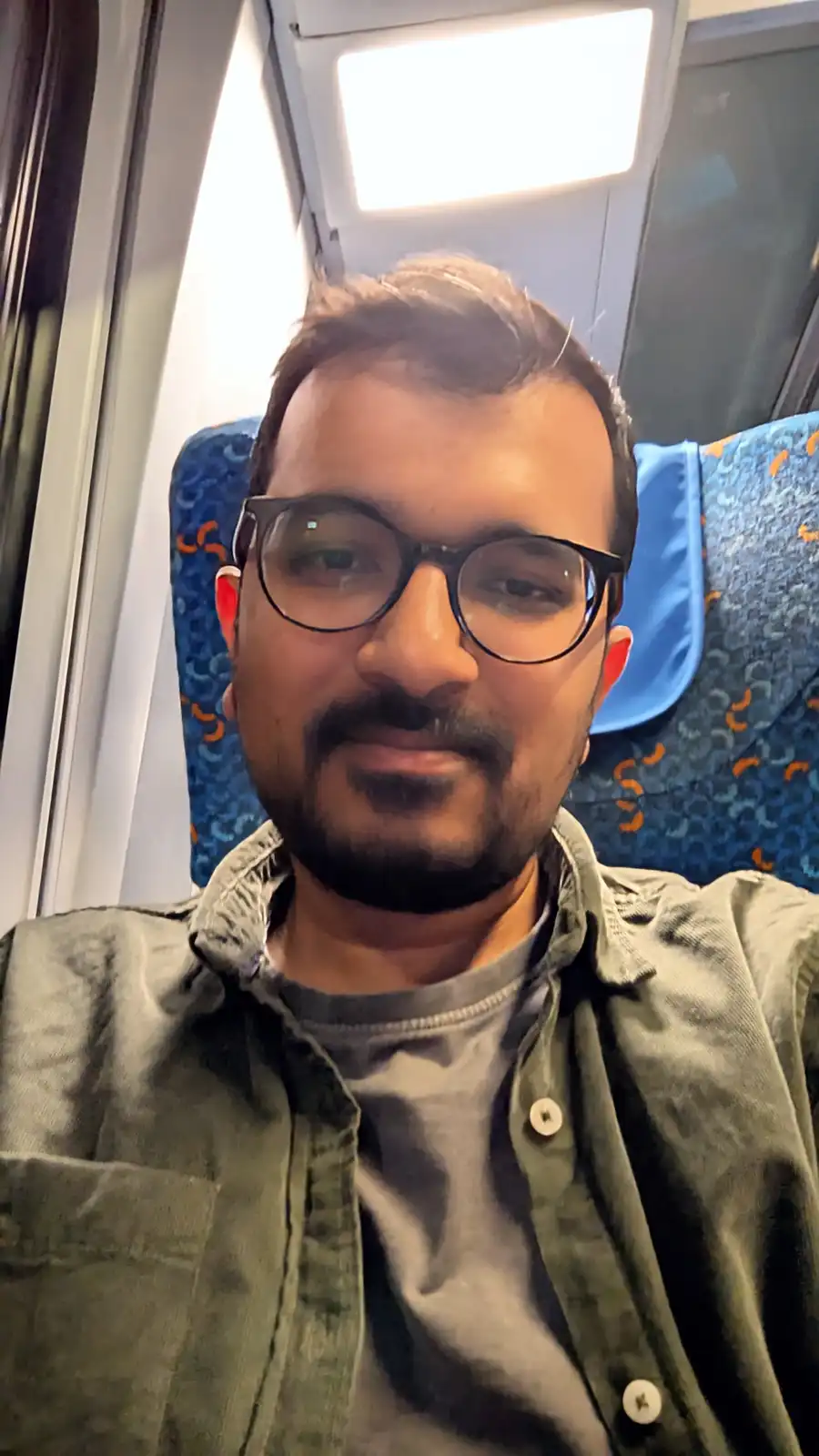A man with glasses and a beard sits in a train seat with blue patterned upholstery, looking directly at the camera with a…
