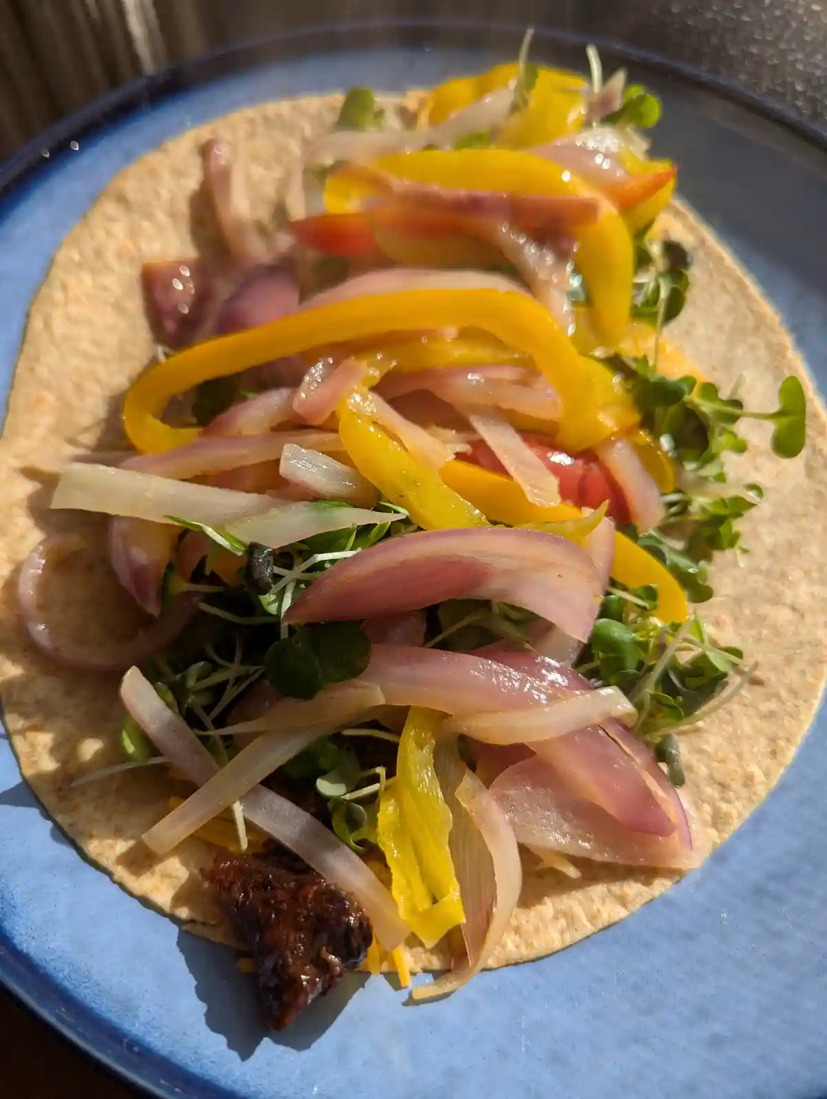 A close-up photo shows a single open-faced taco on a blue plate, filled with yellow bell peppers, sliced red onions, green…