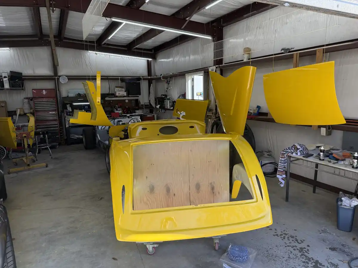 A bright yellow car body in a garage has its rear hatch open, revealing a wooden panel where the back seat should be. Other…