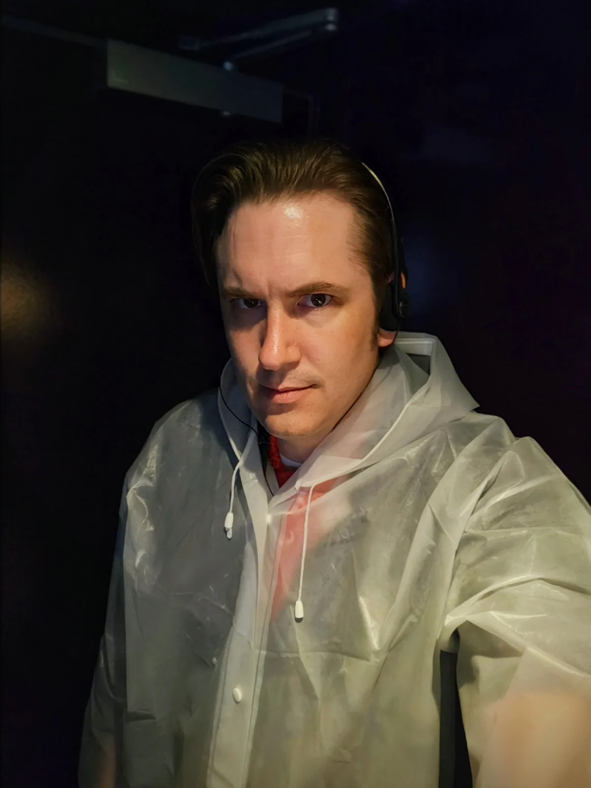 A man wearing a clear plastic rain poncho and headphones looks directly at the camera with a serious, slightly annoyed…