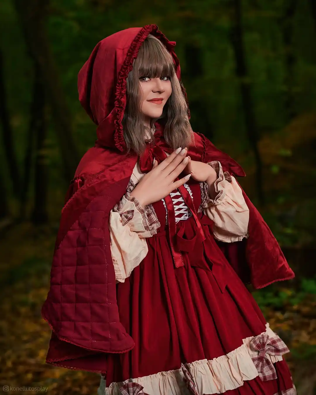 A person in a detailed red hooded cloak and dress stands in a forest, hands clasped near their chest with a gentle smile…