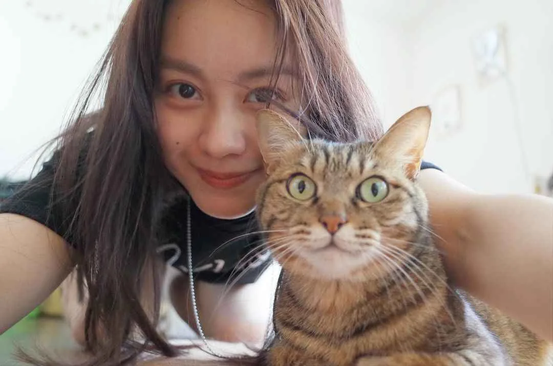 A young woman with long dark hair smiles at the camera while holding a tabby cat that stares directly forward with wide…
