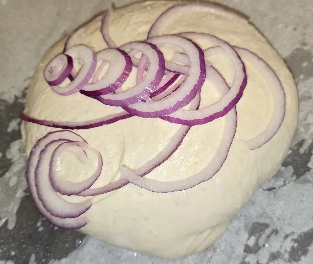 A round ball of unbaked dough is topped with decorative, spiraling slices of red onion arranged to resemble a face or…
