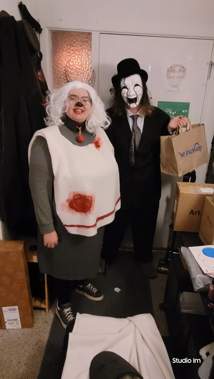 Two people in costumes pose indoors: one wears a white wig, glasses, a fake black nose, and a white vest with red stains…