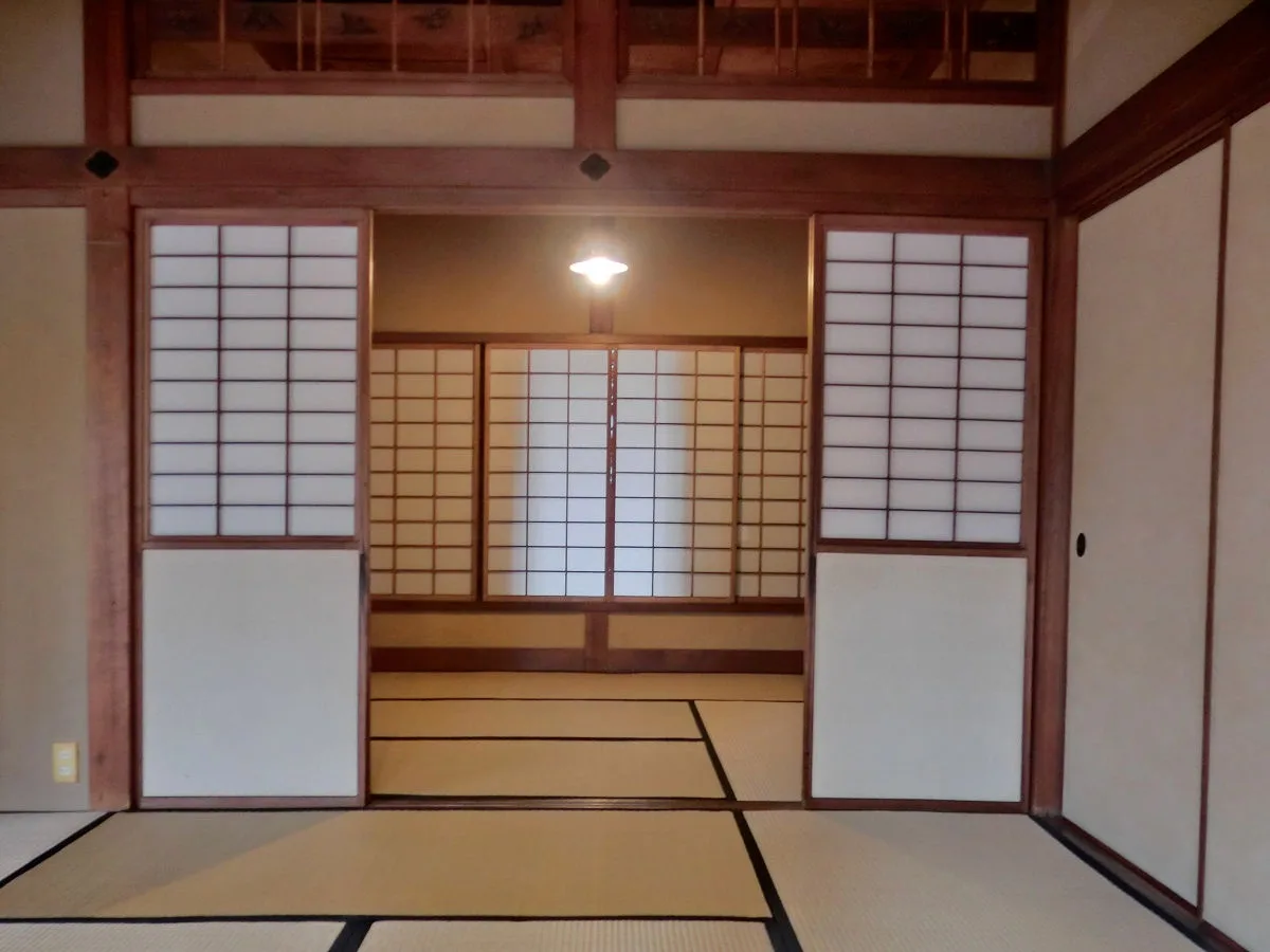 An empty traditional Japanese room with tatami mats, wooden sliding shoji doors, and a single ceiling light. The overlaid…