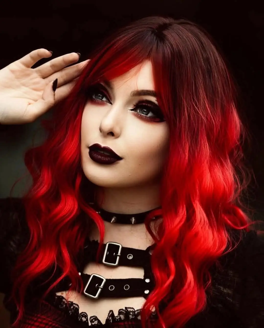 A person with long, vibrant red hair and dark gothic makeup, wearing a black studded choker and a black leather harness top…