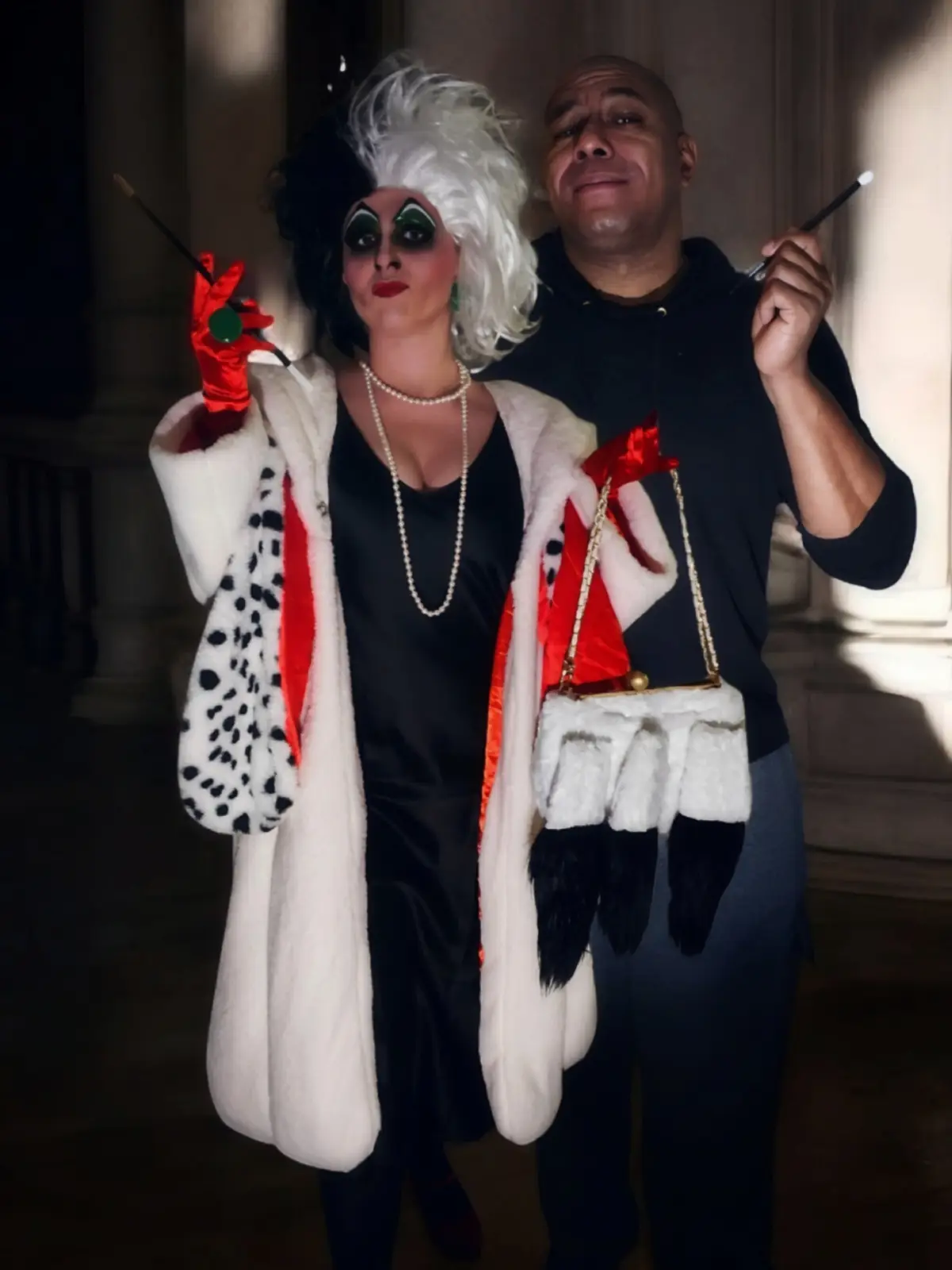 A person dressed as Cruella de Vil, wearing a white fur coat with black spots, red gloves, and dramatic makeup, stands next…