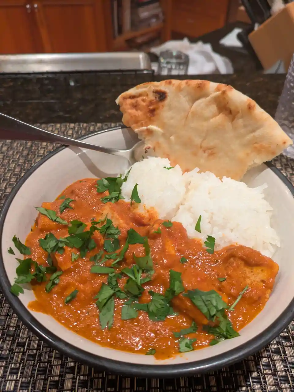 A close-up photo of a bowl of Indian-style chicken curry with white rice and a piece of naan bread, garnished with chopped…