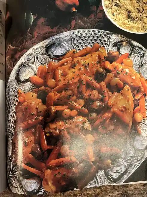 A photo of a cookbook page showing a plated dish of chicken with carrots and chickpeas on an ornate silver platter, with a…