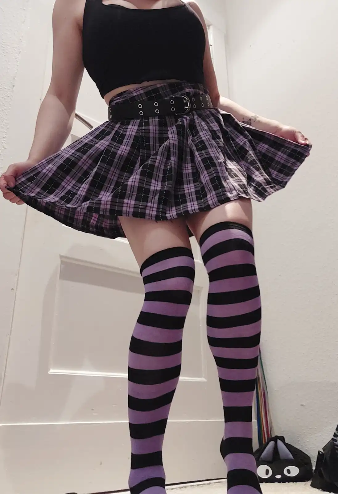 A person wearing a black crop top, a purple and black plaid pleated skirt with a black belt, and purple and black striped…