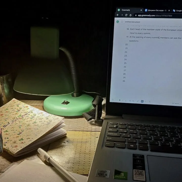 A dimly lit desk setup with a green desk lamp, a floral notebook, and a pen. A laptop screen displays a Grammarly document…