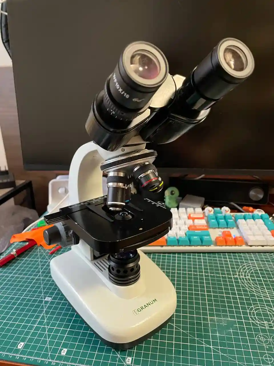A white and black Granum brand stereo microscope sits on a green cutting mat, with a colorful mechanical keyboard and a dark…