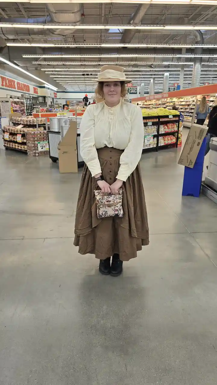 A person dressed in vintage-style clothing, including a cream blouse, brown tiered skirt, and hat, stands in a modern…